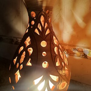 May include: A handcrafted, brown ceramic vase with intricate cut-out patterns. The vase is illuminated from within, casting warm, orange light through the decorative openings. The design features floral and geometric shapes.