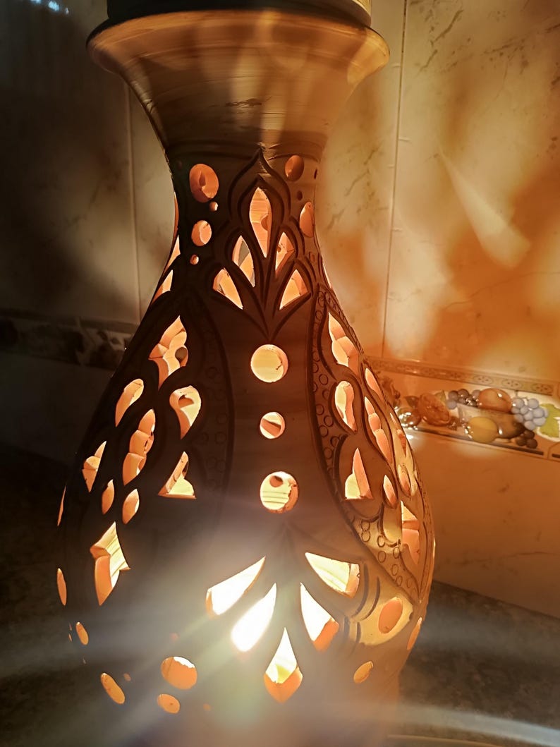 May include: A handcrafted, brown ceramic vase with intricate cut-out patterns. The vase is illuminated from within, casting warm, orange light through the decorative openings. The design features floral and geometric shapes.