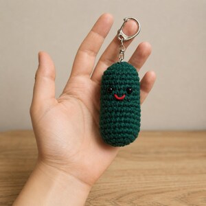 May include: A handmade, dark green crochet pickle keyring with a smiling face. The pickle has black eyes and a red stitched mouth. The keyring is attached to a silver-coloured metal clasp.