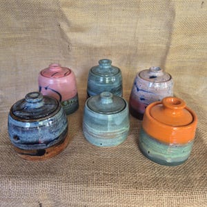 May include: Six small ceramic jars with matching lids, each featuring a unique glaze finish. The collection includes colours such as pink, blue, orange, and multi-toned earth shades. Each jar has a cylindrical body and a small knob on the lid.