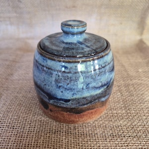 May include: A ceramic lidded jar featuring a cylindrical body with a rounded top. The glaze transitions from a deep terracotta brown at the base to a mottled blue and dark navy finish on the upper section and the matching lid with a knob handle.