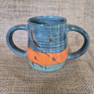 May include: A handmade ceramic mug with two handles. The mug features a teal blue upper section, an orange band in the middle, and a teal green base. Brown streaks and drips add detail. The mug is on a burlap surface.