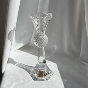 May include: A clear crystal candlestick holder with a fluted top and a textured sphere in the middle. The base is a faceted shape. A small gold label is affixed to the base.