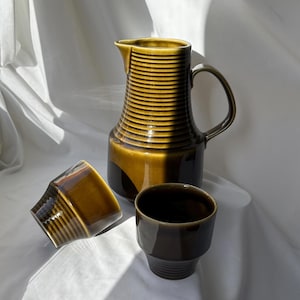 May include: A vintage ceramic pitcher and two matching cups in shades of brown and olive green. The pitcher has a ribbed design and a handle. The cups are also ribbed. The set is displayed on a white surface.