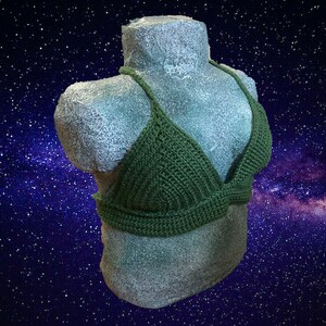 May include: A green crocheted halter top with a triangular shape. The top is displayed on a mannequin against a dark blue background with white stars.