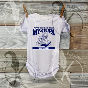 Puede incluir: Body de bebé blanco con un gráfico azul. El gráfico presenta el texto "Pappa weet baie, maar My-Oupa weet alles!" encima de una ilustración de un abuelo leyendo un libro a un niño. El body está colgado en un tendedero.