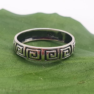 Könnte beinhalten: Ein silberner Bandring mit einem geometrischen Muster um die gesamte Band. Der Ring liegt auf einem grünen Blatt.