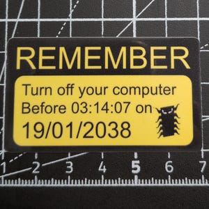 May include: A rectangular sticker with a black border and yellow center. The text reads REMEMBER Turn off your computer Before 03:14:07 on 19/01/2038. A small black insect graphic is positioned to the right of the date. The sticker is placed on a black cutting mat with white grid lines showing measurements in inches.