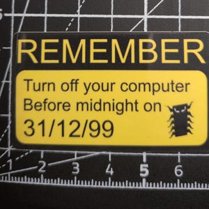 Op de afbeelding: Een rechthoekige sticker met een zwarte rand en een geel midden. De tekst luidt REMEMBER Turn off your computer Before midnight on 31/12/99. Een zwart insect-icoon staat rechts. Het ligt op een zwarte snijmat met witte centimeter markeringen.