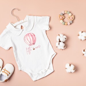 May include: White baby onesie with a pink hot air balloon and the text "little one" printed on it. The onesie is on a pink background with cotton balls and a wooden toy.