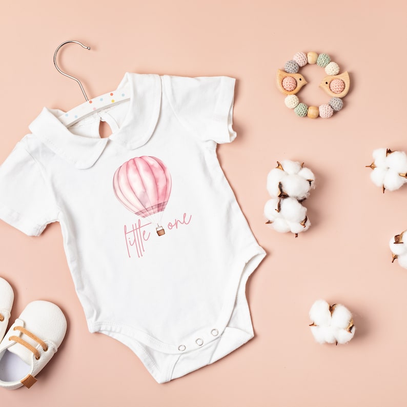 May include: White baby onesie with a pink hot air balloon and the text "little one" printed on it. The onesie is on a pink background with cotton balls and a wooden toy.