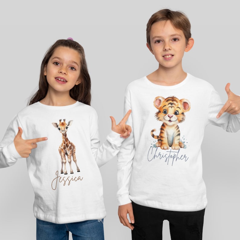 May include: Two children wearing white long-sleeved shirts with animal illustrations and their names printed below. The girl's shirt features a giraffe and the text "Jessica". The boy's shirt features a tiger and the text "Christopher".