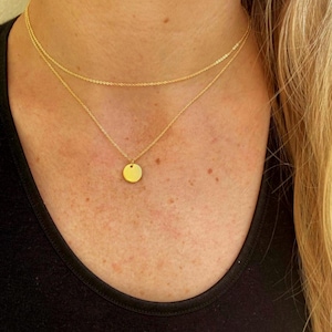 Könnte beinhalten: Zweiteiliges goldfarbenes Halsketten-Set. Die obere Kette ist filigran, während die untere Kette einen kleinen, runden Anhänger hat. Die Halskette wird über einem schwarzen Oberteil getragen, was den Glanz des Schmucks hervorhebt.