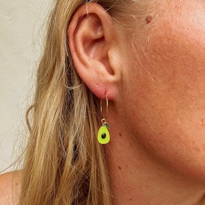 Può includere: Orecchini a cerchio dorati con un ciondolo a forma di avocado verde brillante. L'avocado ha un seme nero e un piccolo dettaglio di foglia verde. Gli orecchini sono indossati da una persona con i capelli biondi.