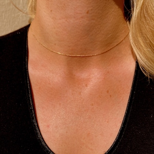 Könnte beinhalten: Eine zarte goldene Halskette, die um den Hals einer Person liegt. Die Kette besteht aus kleinen, verbundenen Gliedern. Die Person trägt ein schwarzes Oberteil, und ihr blondes Haar ist sichtbar.