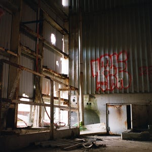 May include: Interior view of an abandoned industrial building with peeling paint and rusted metal structures. Graffiti in red spray paint is visible on the corrugated metal wall. Sunlight streams through holes in the walls and roof, illuminating the decaying space.