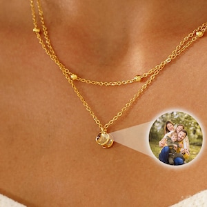 May include: A gold-colored, layered necklace with small beaded accents. The necklace has a small, round pendant with a photo of a family. The necklace is worn on a person's neck.