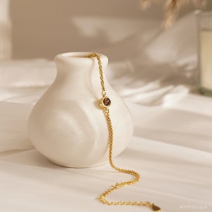 May include: A delicate gold chain bracelet with a single brown gemstone accent. The bracelet is displayed on a white ceramic vase.