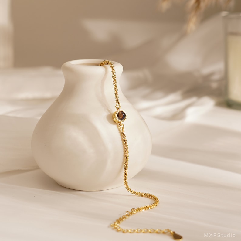 May include: A delicate gold chain bracelet with a single brown gemstone accent. The bracelet is displayed on a white ceramic vase.