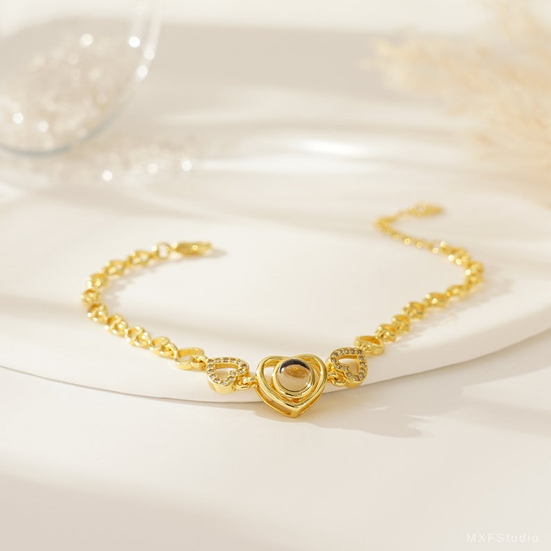 Puede incluir: Un brazalete de cadena dorada con un colgante en forma de coraz&oacute;n. El colgante tiene una peque&ntilde;a ventana redonda en el centro.