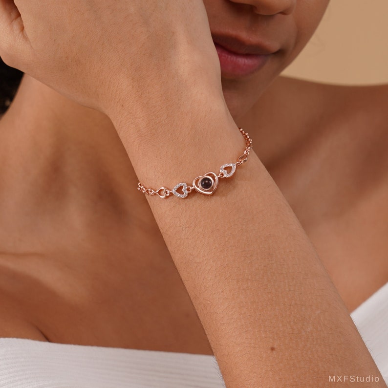 Puede incluir: Un brazalete de oro rosa con un dije en forma de coraz&oacute;n que proyecta un mensaje cuando se ve desde un &aacute;ngulo determinado. El brazalete est&aacute; adornado con dijes m&aacute;s peque&ntilde;os en forma de coraz&oacute;n y piedras de circonita c&uacute;bica transparentes.