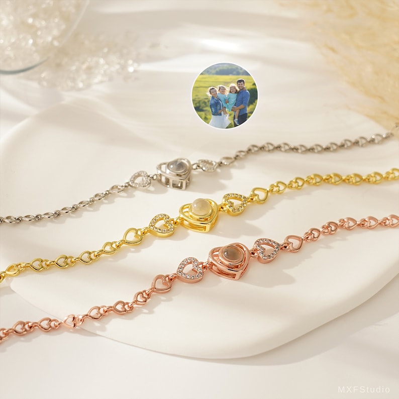 Puede incluir: Tres pulseras de plata, oro y oro rosa con un dije en forma de coraz&oacute;n que proyecta una foto cuando miras a trav&eacute;s del centro. Cada pulsera tiene una cadena de peque&ntilde;os corazones.