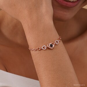 Puede incluir: Una pulsera de oro rosa con un dije en forma de coraz&oacute;n que proyecta un mensaje cuando se ve desde un &aacute;ngulo determinado. La pulsera est&aacute; adornada con dijes m&aacute;s peque&ntilde;os en forma de coraz&oacute;n y piedras de circonita c&uacute;bica transparentes.