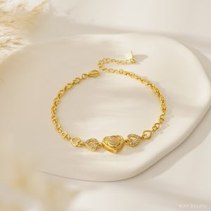 Puede incluir: Un brazalete dorado con un dije en forma de coraz&oacute;n que presenta una piedra transparente en el centro. El brazalete est&aacute; hecho de una cadena delicada con peque&ntilde;os eslabones en forma de coraz&oacute;n.