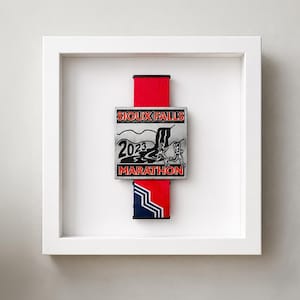 May include: A framed marathon medal from the Sioux Falls 2023 Marathon. The medal is silver with black text and a red and blue ribbon. The frame is white.