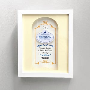 May include: A framed print featuring a cream-colored background with a decorative gold border. The print includes the text "PRESTON KANSAS CITY, MO" and the quote "It takes People to Make the Dream a Reality." The frame is white.