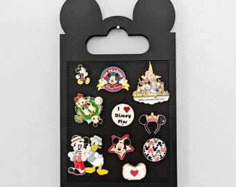 May include: A black display board shaped like Mickey Mouse ears, holding various Disney-themed enamel pins. Pins feature characters like Mickey Mouse, Minnie Mouse, and Donald Duck, along with park-related designs. The pins are colorful and detailed.