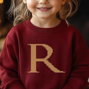 May include: A burgundy sweatshirt featuring a large, gold-colored letter "R" in a knitted style. The sweatshirt is worn by a child. The background is blurred, with hints of warm lighting.