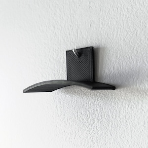 May include: A black, wall-mounted shelf with a curved design. The shelf is attached to a square backplate with a single screw. The shelf is made of a solid material and is designed to hold small items. The shelf is mounted on a white wall.