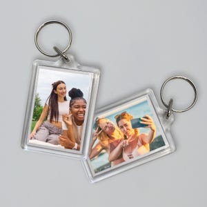 Op de afbeelding: Twee doorzichtige acryl sleutelhangers met zilveren ringen. Elke sleutelhanger bevat een rechthoekige foto. Eén foto toont drie vrouwen die buiten poseren, en de andere toont twee vrouwen die een selfie maken.