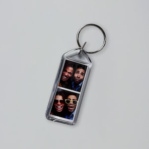 Puede incluir: Llavero rectangular transparente con un anillo de metal plateado. El llavero contiene dos tiras de fotos, cada una con dos personas sonriendo. Una persona lleva gafas de sol amarillas. El fondo es gris claro liso.
