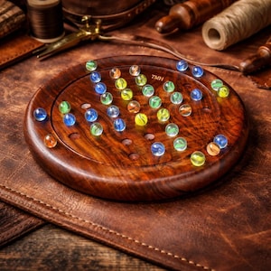 Könnte beinhalten: Ein Solitärspiel aus Holz mit einem kreisförmigen Brett und Löchern, gefüllt mit bunten Glasmurmeln. Das Brett hat eine satte braune Farbe, und die Murmeln sind in verschiedenen Farben, darunter Blau, Grün, Gelb und Orange. Der Text "TMH" ist sichtbar.