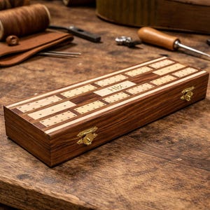 Op de afbeelding: Een rechthoekig houten cribbagebord met een donkerbruine afwerking en licht houten inlegwerk. Het bord heeft messingkleurige sluitingen en de initialen "HBC" zijn op de bovenkant gegraveerd. Het spelbord heeft rijen gaten om te prikken.