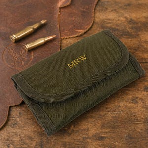 May include: An olive green canvas case with the initials "MRW" in gold lettering. Two brass bullets are positioned near the case on a brown leather surface. The background is a wooden table.
