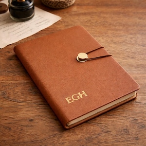 Op de afbeelding: Een bruin leren notitieboek met de gouden initialen "EGH". Het notitieboek heeft een gouden sluiting en een bruine koordsluiting. Het gesloten notitieboek ligt op een houten oppervlak.