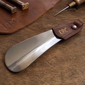 May include: A polished metal shoe horn with a brown leather handle. The handle is embossed with the letters "ERC" in gold. The shoe horn is on a wooden surface.
