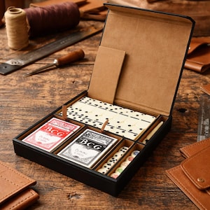 May include: A black leather games set with a hinged lid, containing dominoes, playing cards, and dice. The playing cards are labelled "BCG Playing Cards". The set is displayed on a wooden surface, with leather accessories in the background.