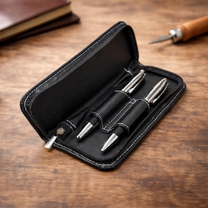 Puede incluir: Un estuche de cuero negro con costuras blancas, abierto para revelar dos bolígrafos plateados y negros. El estuche está sobre una superficie de madera, con un libro encuadernado en cuero y una herramienta de artesanía en el fondo.