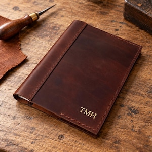 Puede incluir: Una funda de cuaderno de cuero marrón oscuro con una costura vertical en el lado izquierdo y las iniciales TMH en relieve de color dorado en la esquina inferior derecha. La funda tiene una textura mate suave con grano de cuero natural visible.
