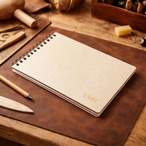 May include: A light-coloured wooden notebook with a spiral binding, resting on a brown leather surface. The initials "TMH" are printed in gold on the bottom right corner. A pencil and other craft tools are visible.