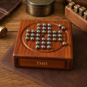 Könnte beinhalten: Ein quadratisches Solitaire-Spiel aus Holz mit einer kreisförmigen, vertieften Spielfläche, auf der zahlreiche silberne Metallkugeln in einem Kreuzmuster angeordnet sind. Die Vorderseite des Holzsockels ist mit den Großbuchstaben TMH graviert.