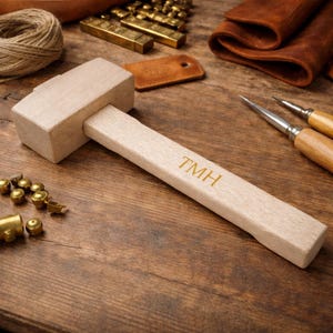 Puede incluir: Un mazo de madera clara con las iniciales "TMH" grabadas en el mango, rodeado de herramientas y suministros de artesanía en cuero. El conjunto incluye herrajes de latón, retales de cuero y un carrete de hilo. Las iniciales son doradas.