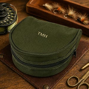 May include: A semi-circular olive green canvas storage case featuring a black zip closure and the embroidered letters TMH in a light gold thread on the top lid. The case has a structured, rounded shape with reinforced piping along the edges.