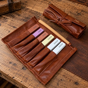 May include: A brown leather roll-up case containing five rectangular sharpening stones with grit labels 120#, 240#, 600#, 1000#, 2000#, and 3000#. The case features a leather tie closure and the embossed letters TMH on the exterior flap.