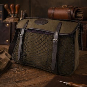 Può includere: Borsa messenger in tela verde oliva con patta e cinturini in pelle marrone scuro. La borsa presenta una tasca frontale in rete e una toppa in pelle con testo in rilievo. La borsa è su una superficie di legno.