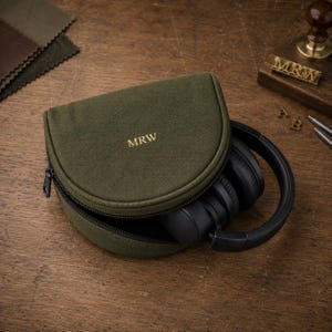 May include: A semi-circular olive green canvas headphone case with a black zipper closure and the letters MRW embroidered in gold thread on the top. Inside the partially open case, a pair of black over-ear headphones are visible.